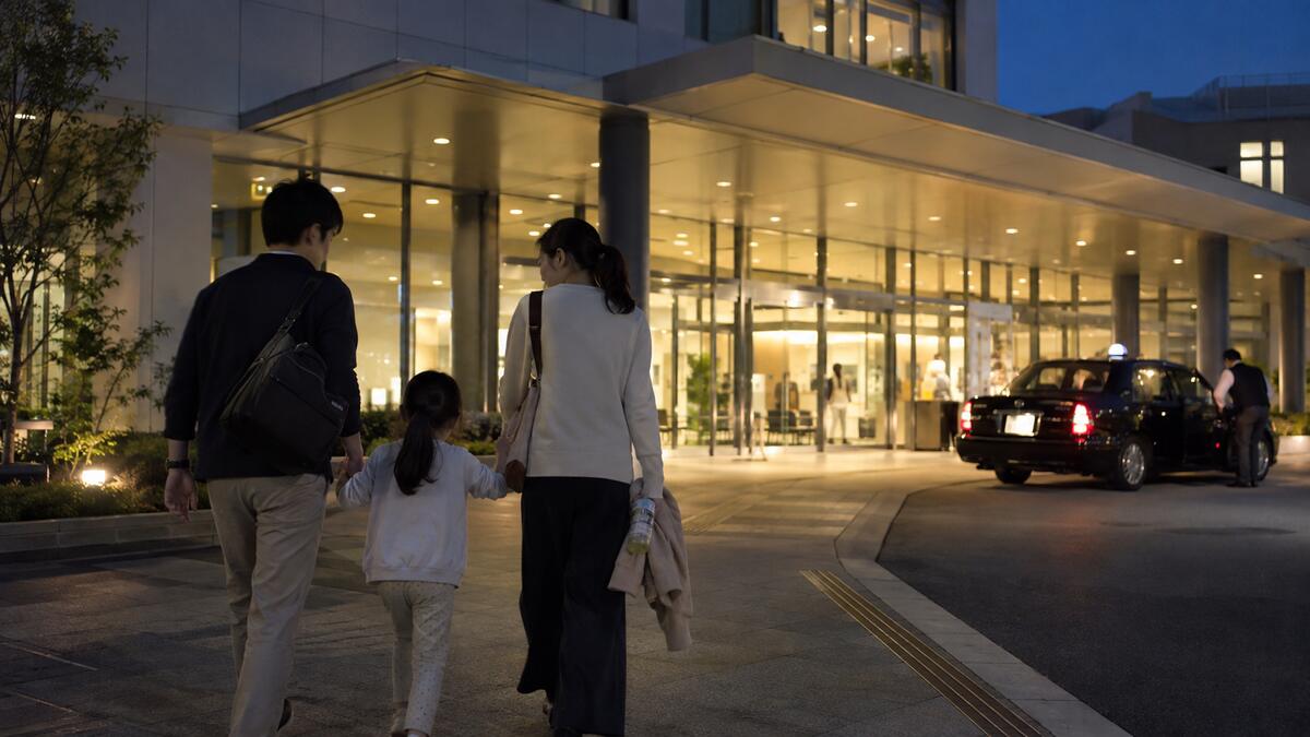 A family walking hand in hand toward a lit clinic entrance at night, with a taxi waiting at the driveway.