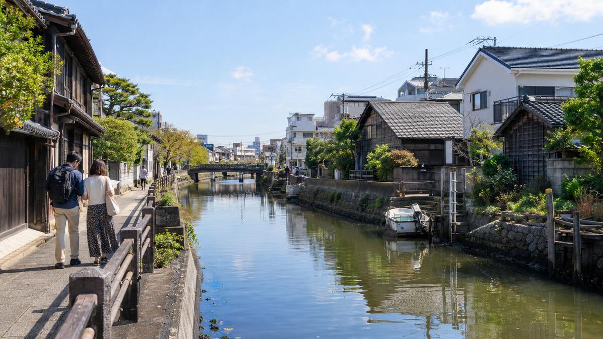 A couple walking beside the Sakaigawa canal near Urayasu Station, past moored boats and old wooden houses.